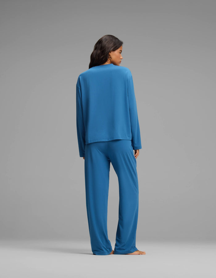 A woman with dark hair stands with her back to the camera against a plain gray background, wearing the Cozy Earth Women’s First Layer Long Sleeve Sleep Tee & Pajama Pant Set in blue.|Color:Anchor