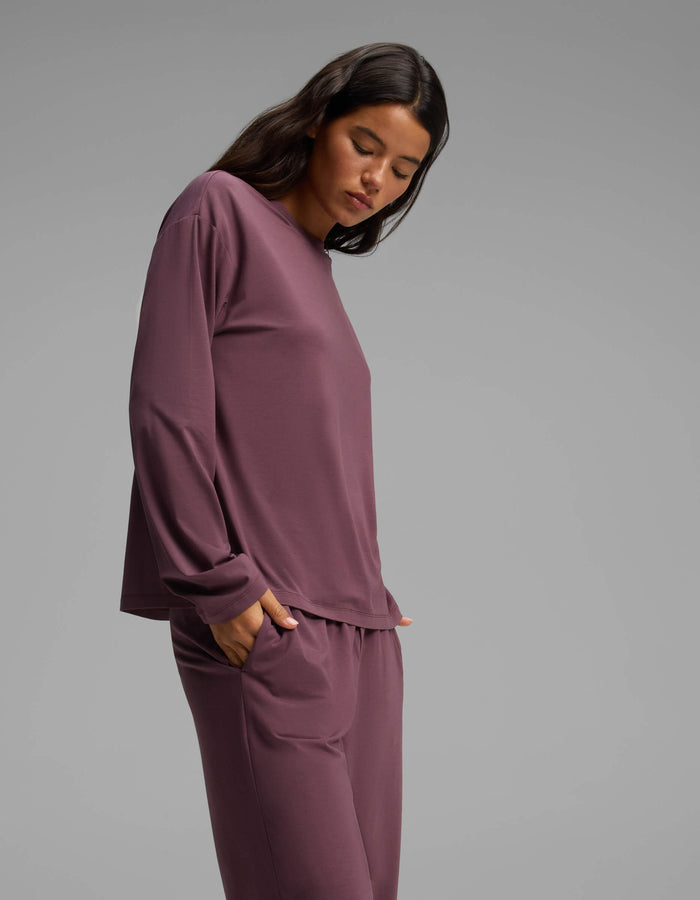 A woman with dark hair stands against a gray backdrop, wearing the Cozy Earth Women’s First Layer Long Sleeve Sleep Tee & Pajama Pant Set in a rich plum shade. She has her hands tucked into the pockets of her comfortable pajama pants and is looking downward.|Color:Fig