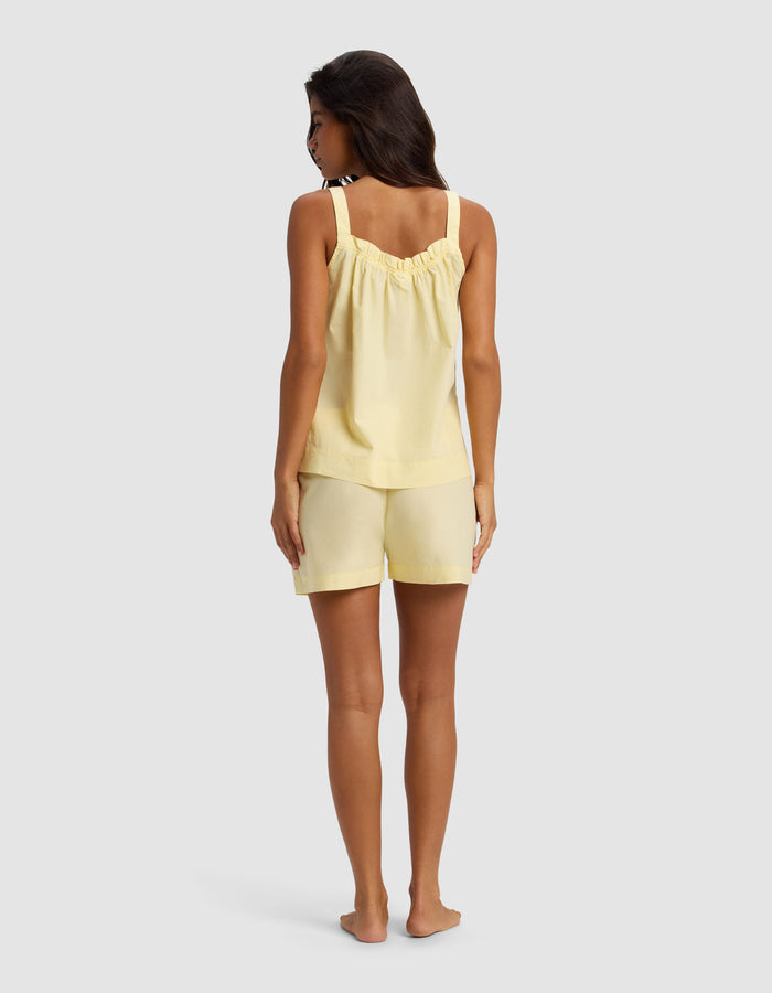 A woman stands barefoot facing away against a light gray background, wearing the Cozy Earth Women's Sutton Tank & Short Pajama Set in pale yellow with ruffled straps.|Color:Meringue