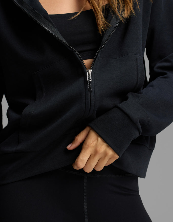 A person wearing the Cozy Earth Women's Versa Full-Zip Hoodie in black, partially unzipped to reveal a black sports bra and part of their hand and upper body, poses against a neutral background.|Color:Jet Black