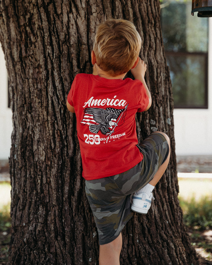 A young boy seen from behind, wearing a red graphic t-shirt and camouflage shorts, climbing a large tree trunk outdoors.